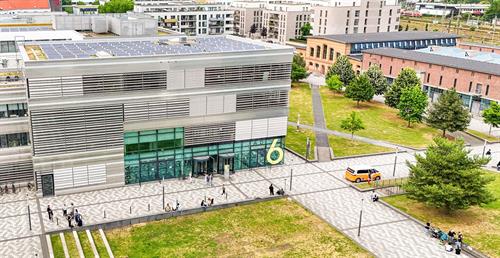 Luftaufnahme des Campus Derendorf der Hochschule Düsseldorf: Im Vordergrund ein modernes Gebäude mit Glasfassade und Solarpaneelen auf dem Dach, davor Studierende auf einem Platz; im Hintergrund weitere Gebäude, Grünflächen und Bahngleise.